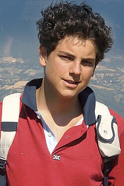 Retrato do Beato Carlo Acutis sorrindo, usando uma camisa polo vermelha e uma mochila, com o Santuário de Assis ao fundo