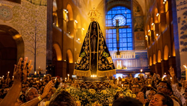 Imagem realista de Nossa Senhora Aparecida iluminada por luz dourada dentro do Santuário Nacional de Aparecida, cercada por fiéis brasileiros em oração, velas acesas e atmosfera espiritual de fé, paz e esperança