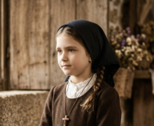 Retrato artístico e realista de uma menina inspirada em Santa Jacinta Marto, com aparência de cerca de 7 a 9 anos de idade. Ela veste roupas camponesas humildes do início do século XX, incluindo um vestido escuro de mangas compridas, um lenço azul-marinho cobrindo a cabeça e um crucifixo de madeira pendurado no pescoço. A menina está de pé, com as mãos unidas à frente do corpo, em uma postura serena e contemplativa. O cenário ao fundo remete a uma casa rústica de pedra e madeira, com iluminação suave vinda de uma janela lateral. A estética da imagem possui tons terrosos e uma textura que lembra fotografias antigas e históricas