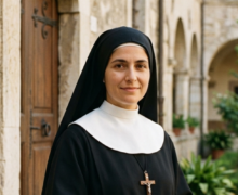 Retrato fotográfico de Santa Maria Domingas Mazzarello representada como uma jovem freira salesiana. Ela sorri suavemente, vestindo um hábito religioso preto tradicional com um colarinho branco (clergyman) e um véu preto. No peito, carrega um crucifixo de madeira simples pendurado por um cordão preto. Ela segura um pequeno livro de orações marrom e um terço entre as mãos. O cenário ao fundo é um pátio interno de um mosteiro de pedra rústica com arcos e janelas antigas, sob iluminação natural diurna